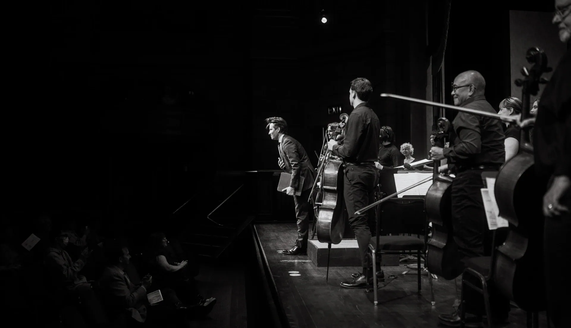 Orchestra musicians stand and sit onstage as seen from the side.