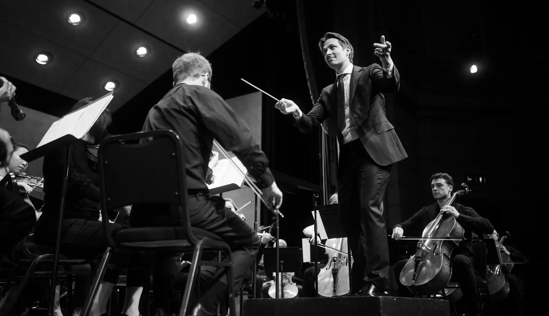 Conductor directs seated orchestra musicians from the front of the stage.