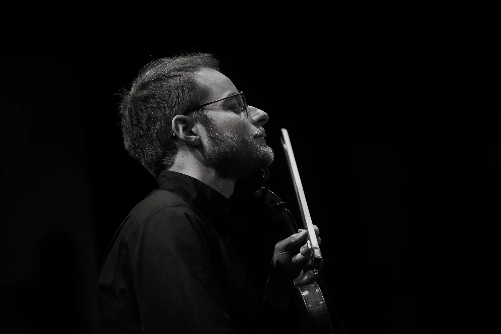 Violinist wearing glasses holds a bow while looking toward the stage.