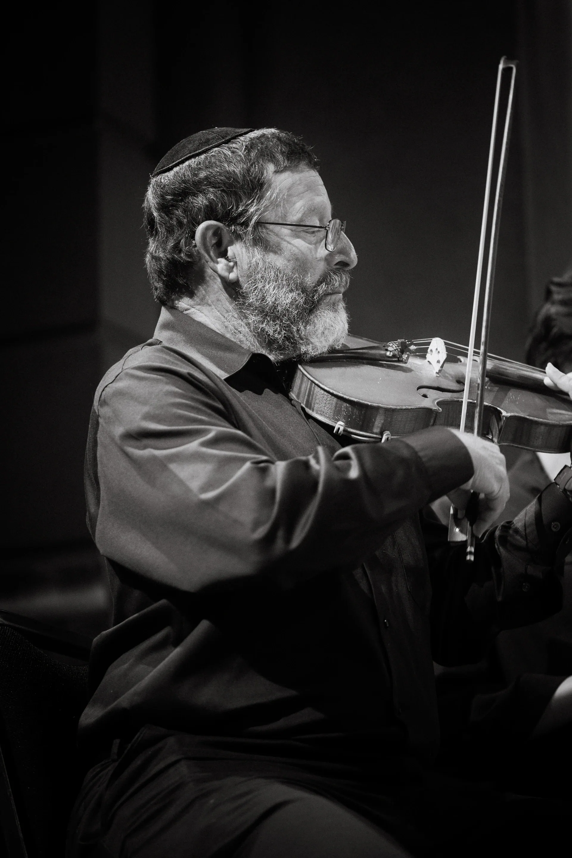 Seated bearded violinist plays beneath soft stage light.