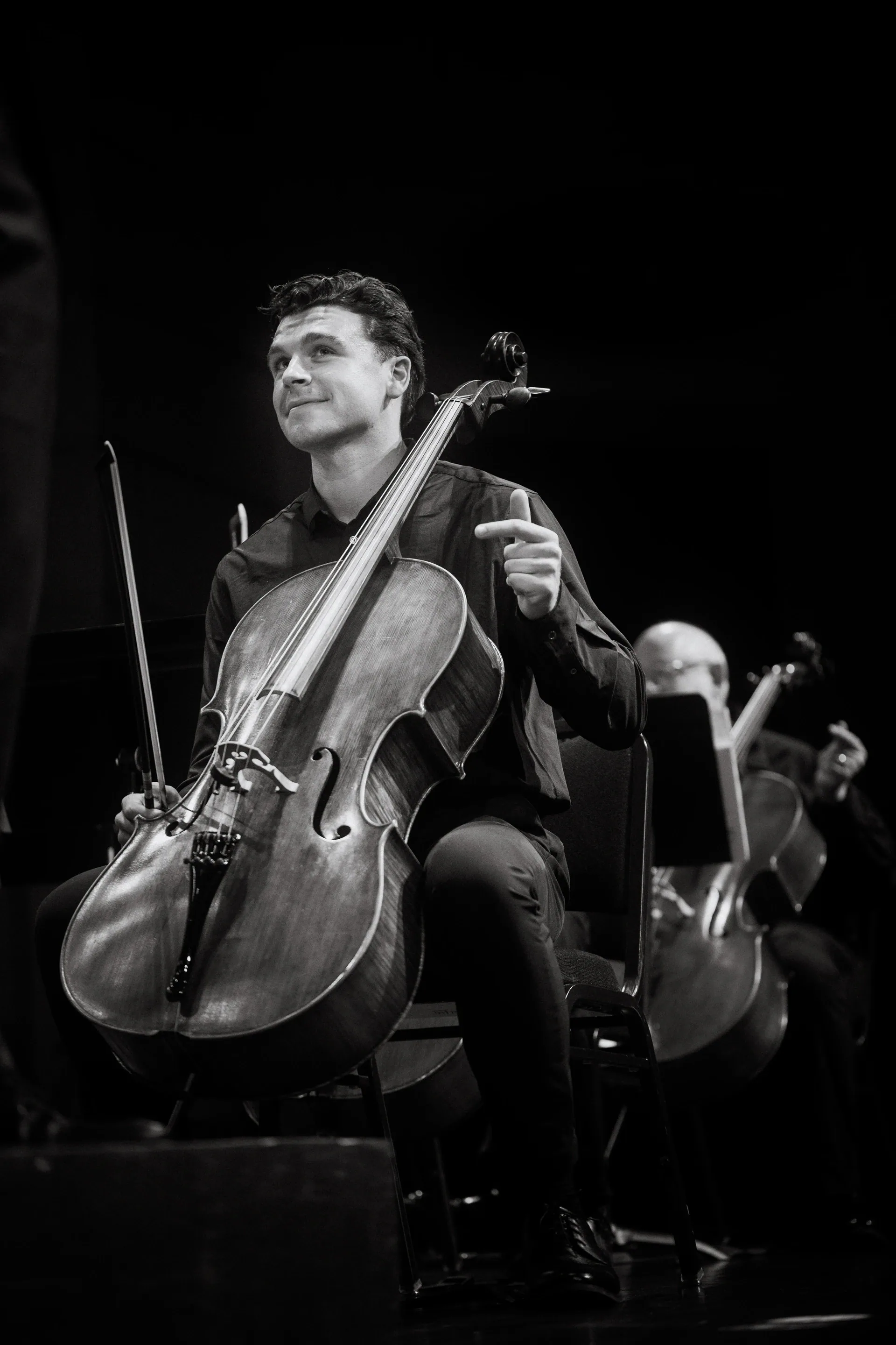 Standing cellist smiles while holding the instrument beside other musicians.