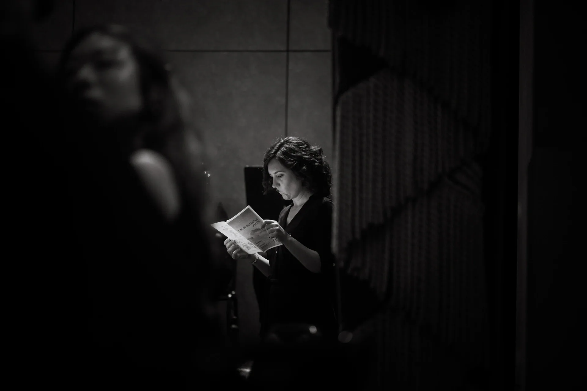 Person stands in dim light reading from a small book or program.