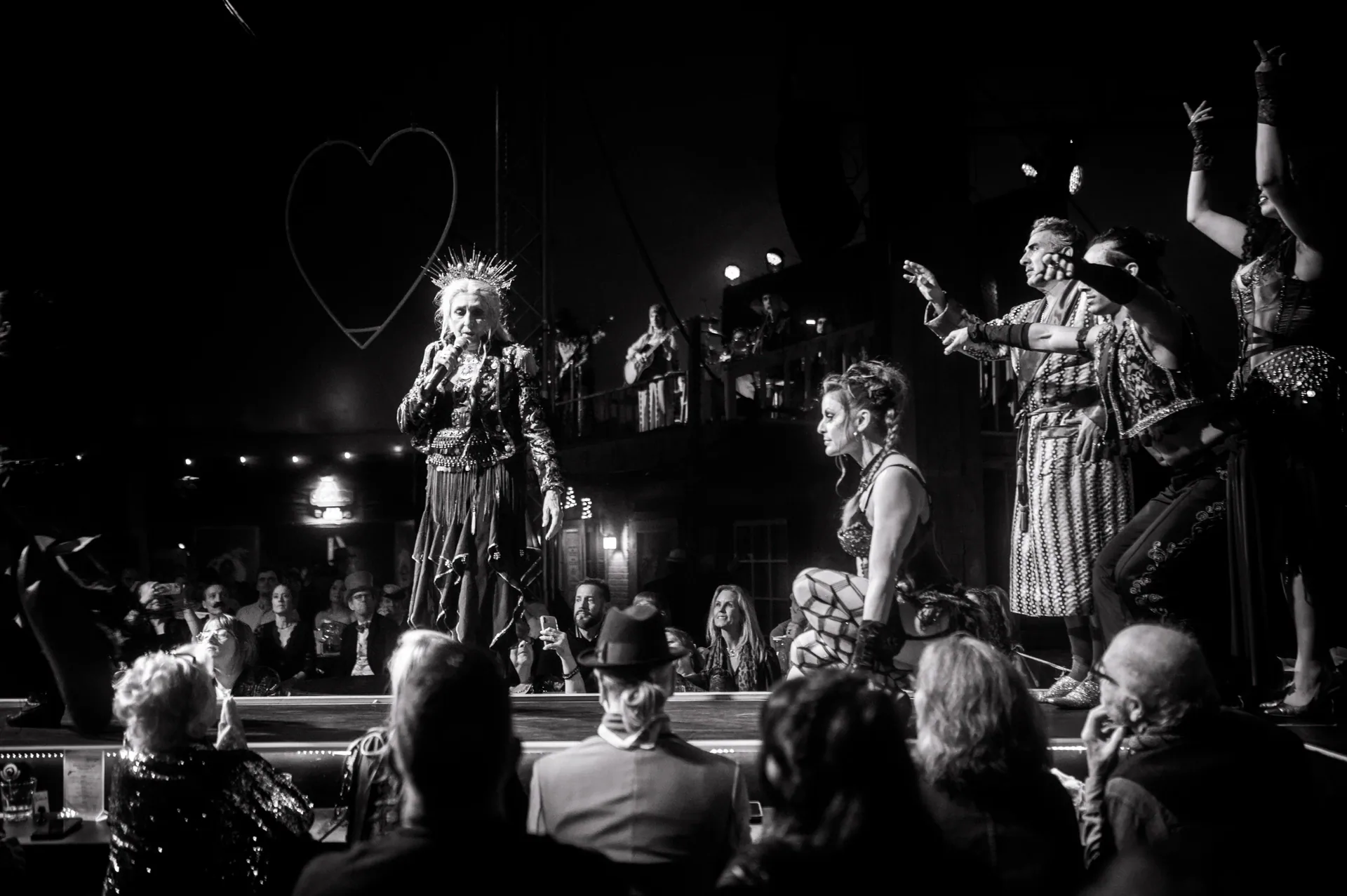 Costumed performers address the audience beside a hanging heart-shaped decoration.