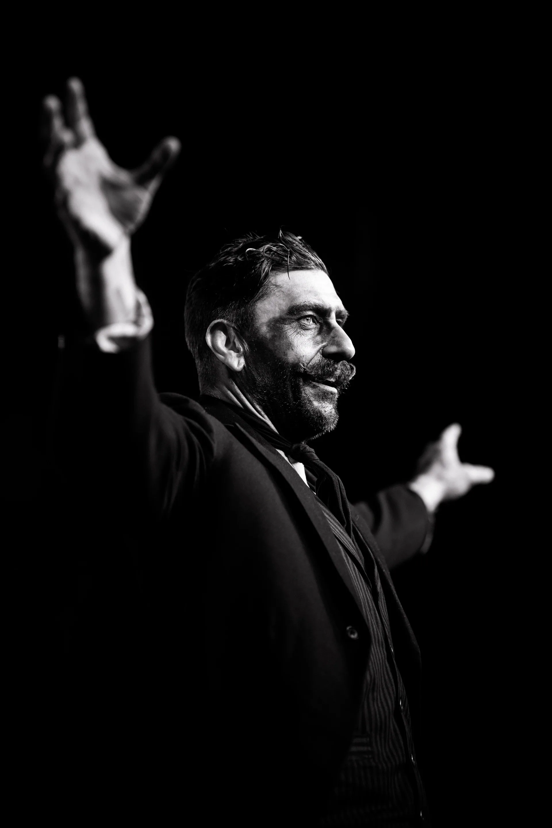 Bearded performer in dark clothing gestures with both arms raised.