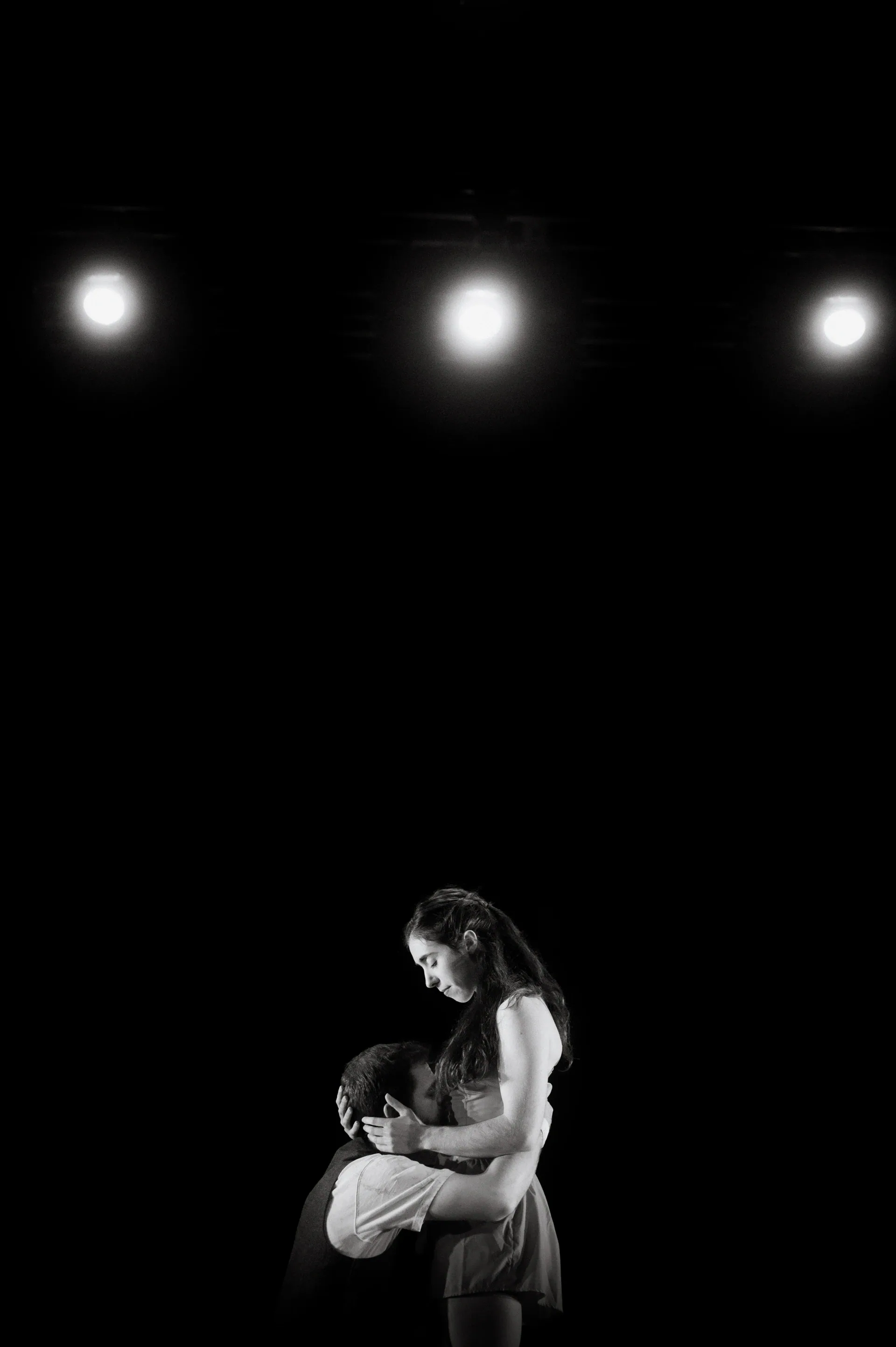 Two performers embrace low on the stage beneath three lights.