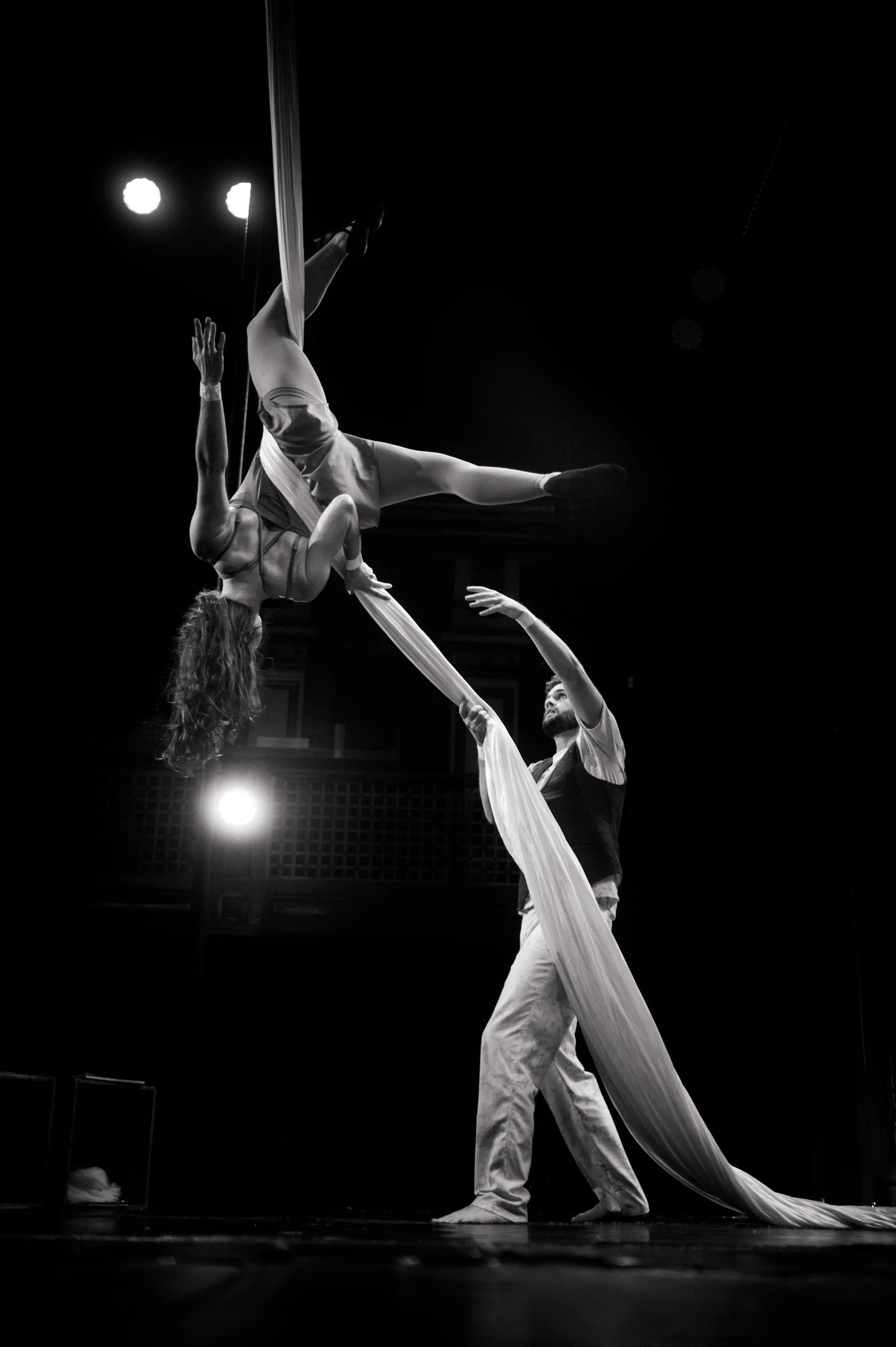 Aerialist reaches from suspended silk toward a standing partner.