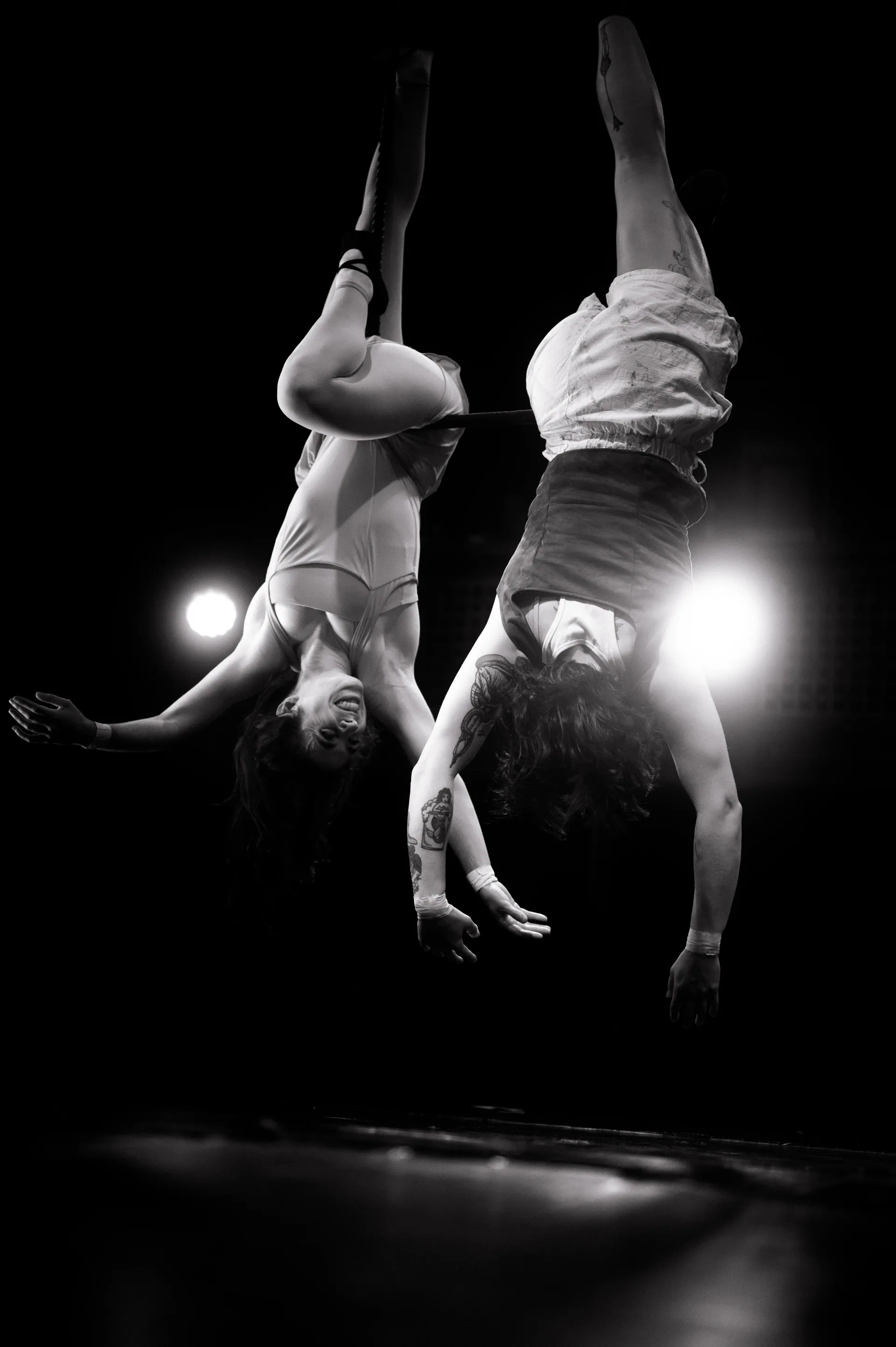 Two aerialists hang upside down side by side beneath stage lights.
