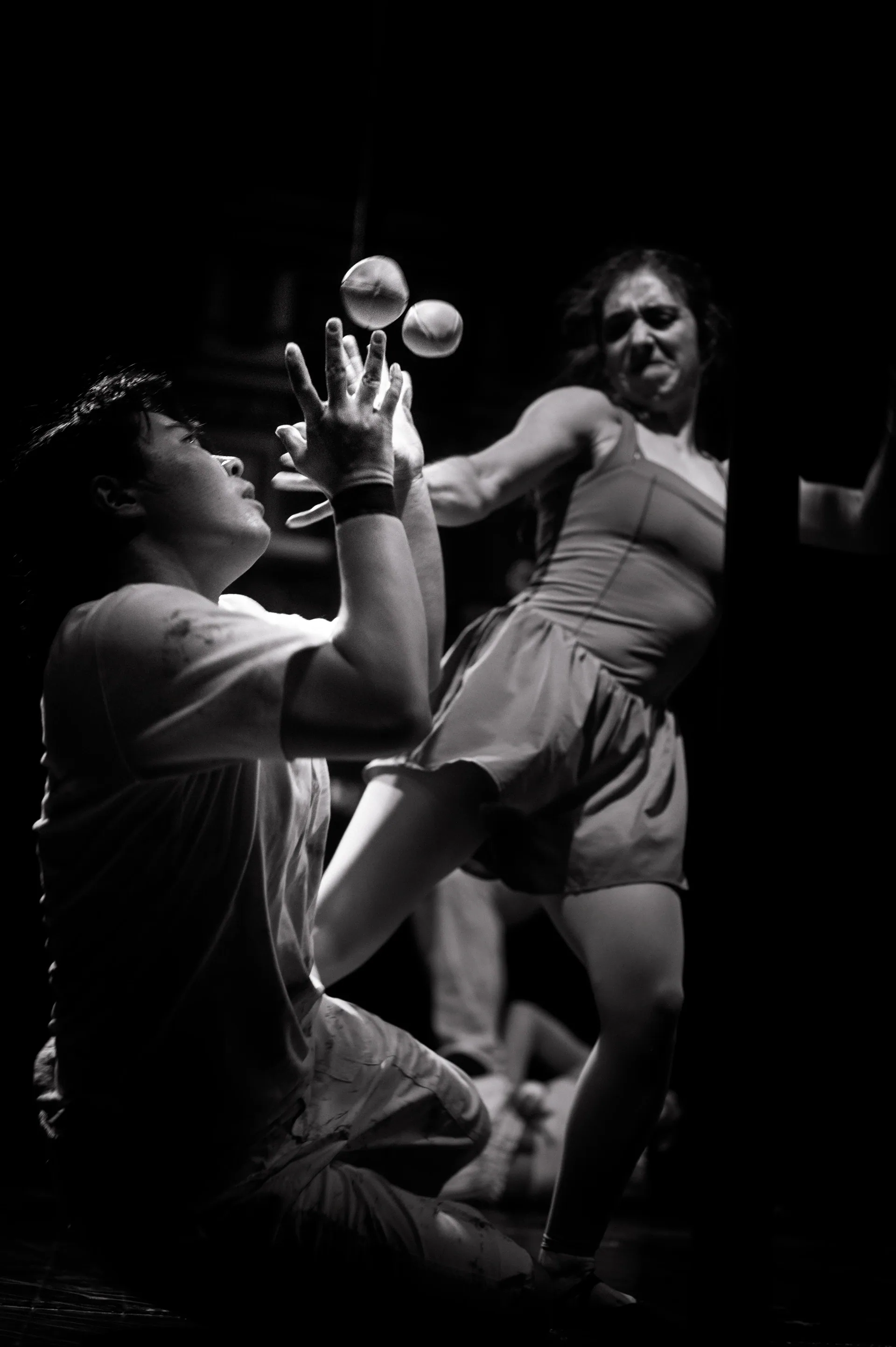 Kneeling performer balances two small translucent balls above the hands while another stands behind.