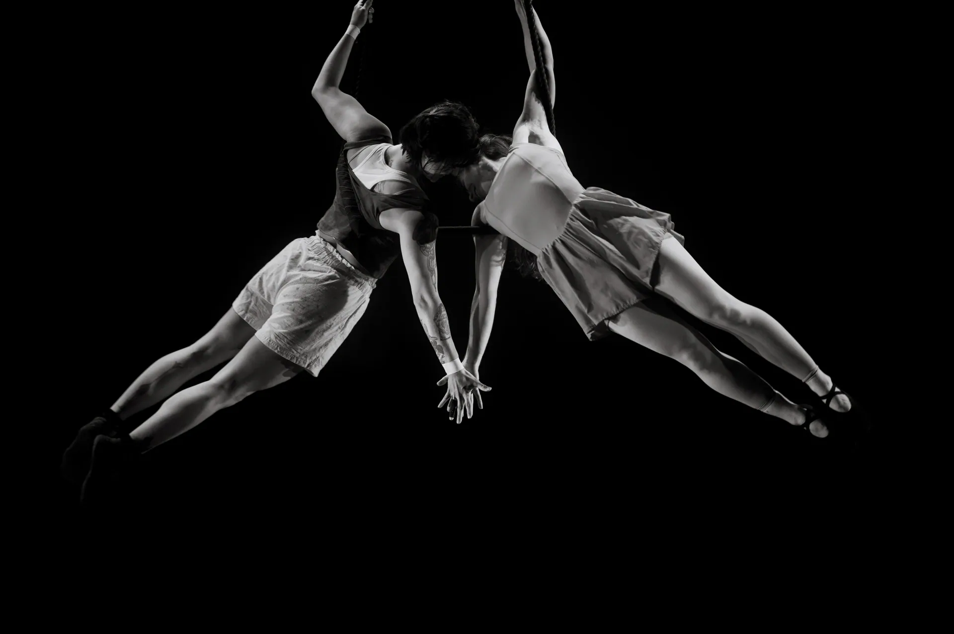 Two aerialists hang horizontally toward each other from separate straps.
