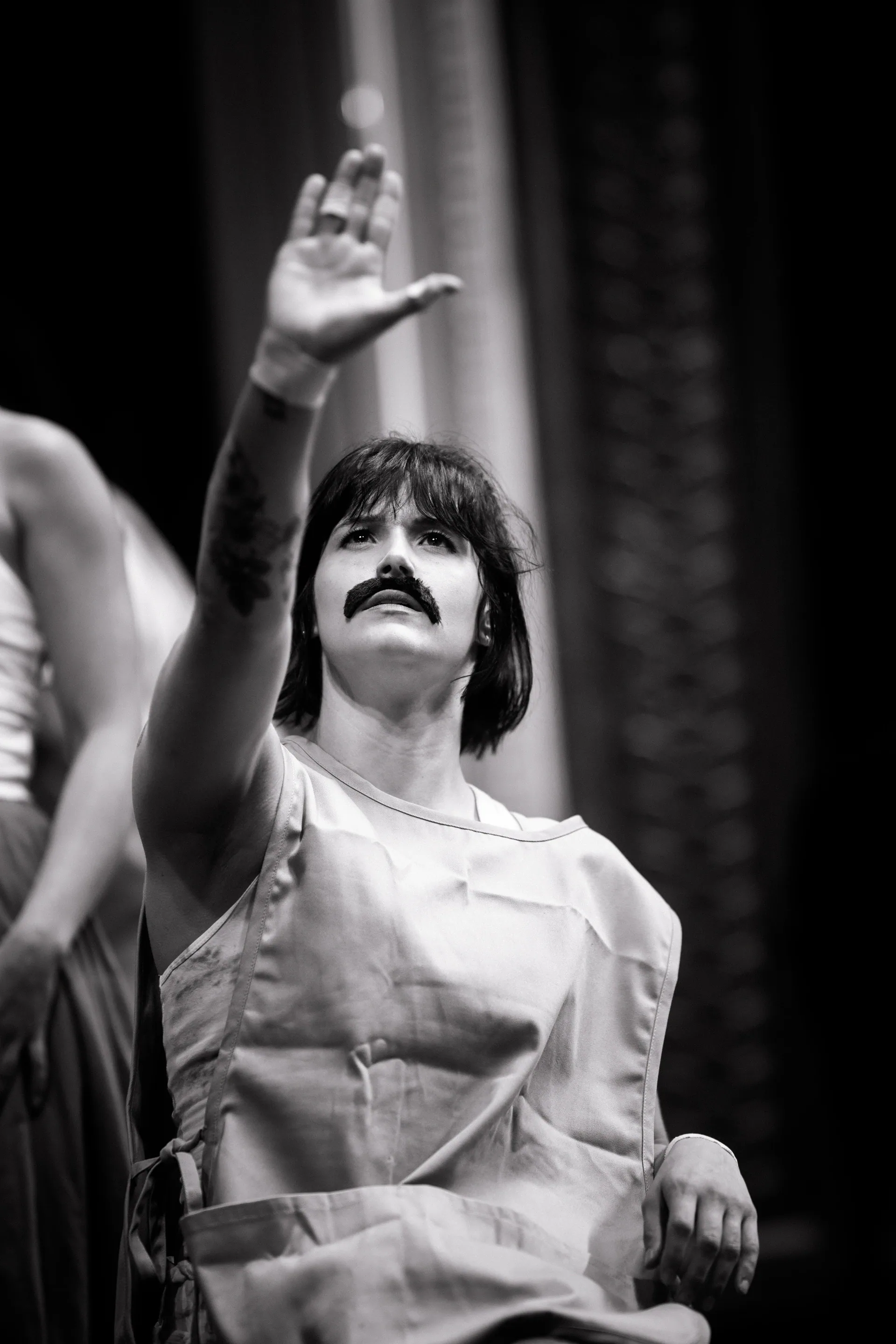 Performer with a painted mustache raises one hand beneath a spotlight.