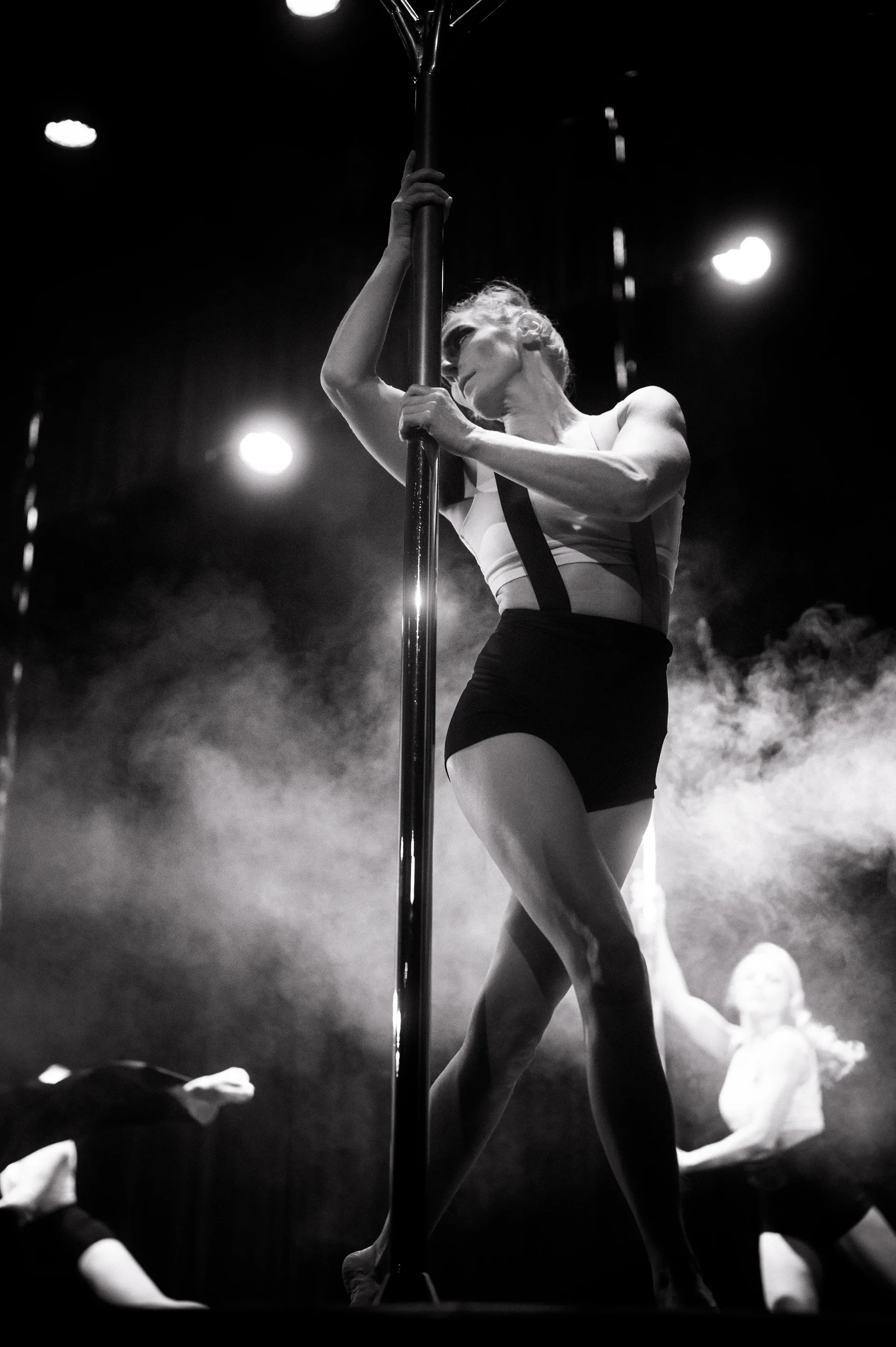 Performer reaches upward while climbing a vertical pole amid stage haze.