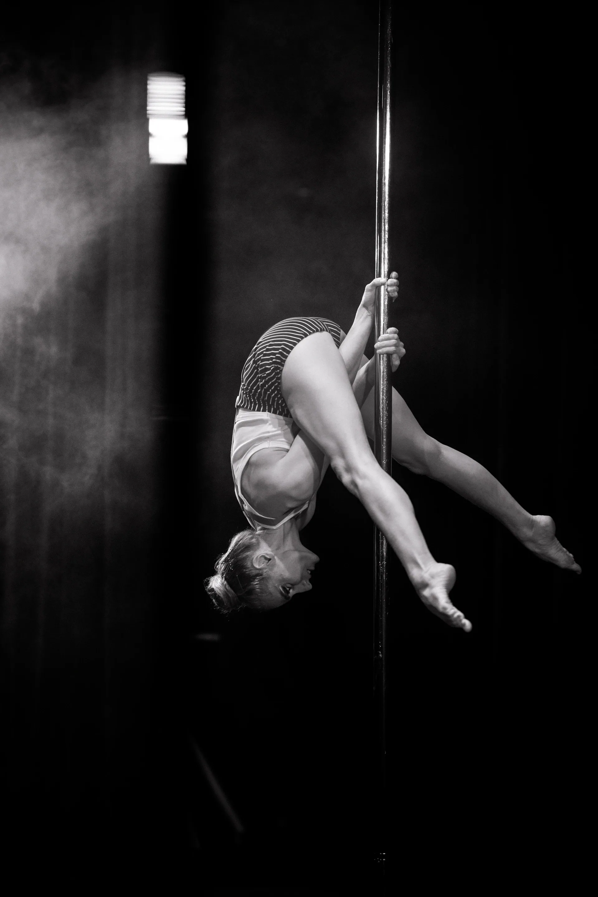 Aerialist hangs upside down from a pole with both legs bent.